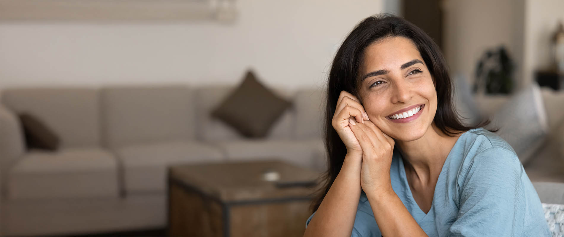 A woman with dark hair smiling at the camera, wearing blue top, sitting on a couch indoors.