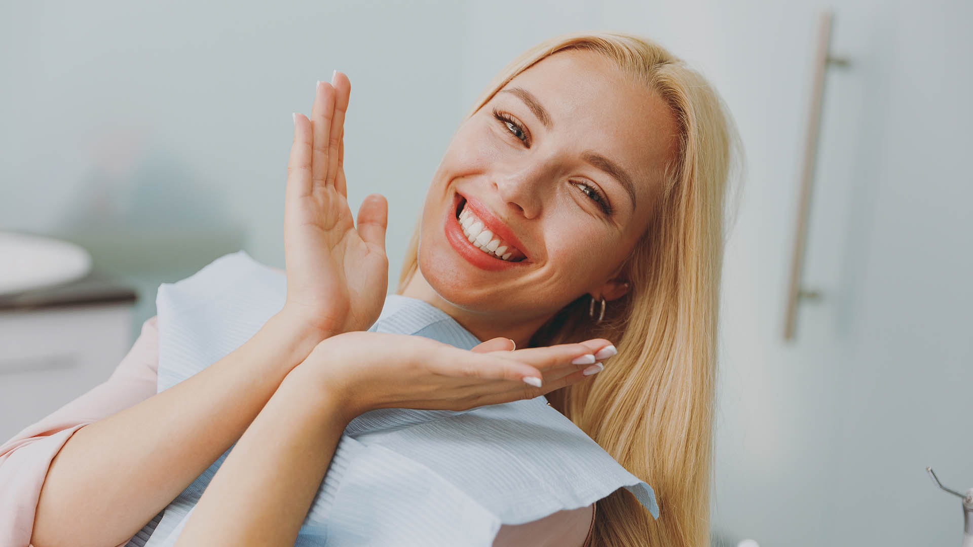 A woman with blonde hair smiling broadly while sitting in a dental chair, holding her hands together in front of her mouth.