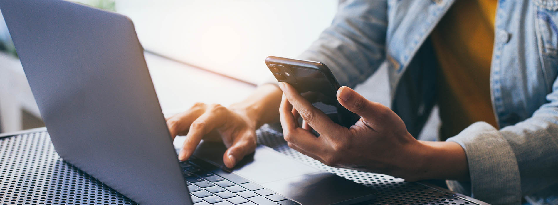 A person using a laptop at a table with a cell phone in hand.