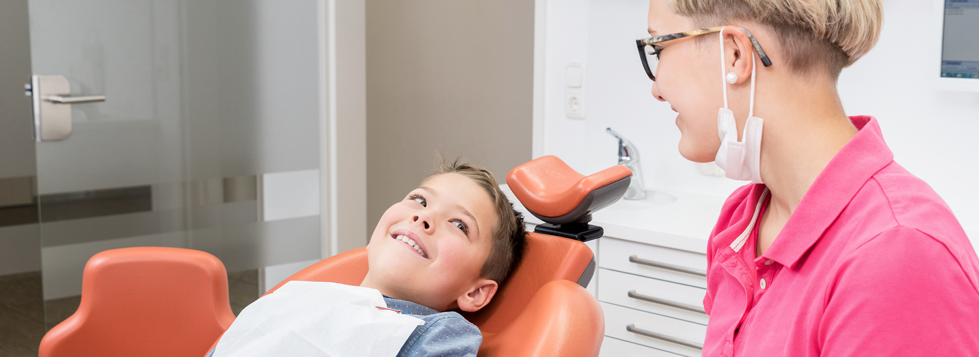 The image shows a dentist and patient in an office setting the dentist is seated at a desk with a computer monitor, while the patient is sitting on a dental chair, smiling and looking towards the camera.